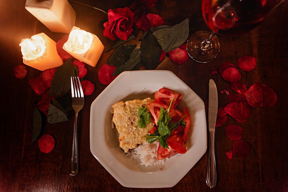 Romantic table setting with food, candles, and red petals for a cozy dinner.